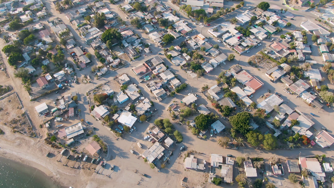 High drone view of Aldofo Lopez Mateos with clustered rooftops, branching dirt streets, small yards, dry open lots, trees, and no visible people.