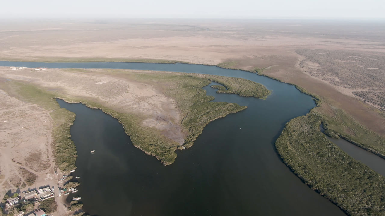 High drone frame over Aldofo Lopez Mateos, Baja California Sur, showing a curved water channel, green scrub islands, sandy flats, a distant water strip, and no visible people.