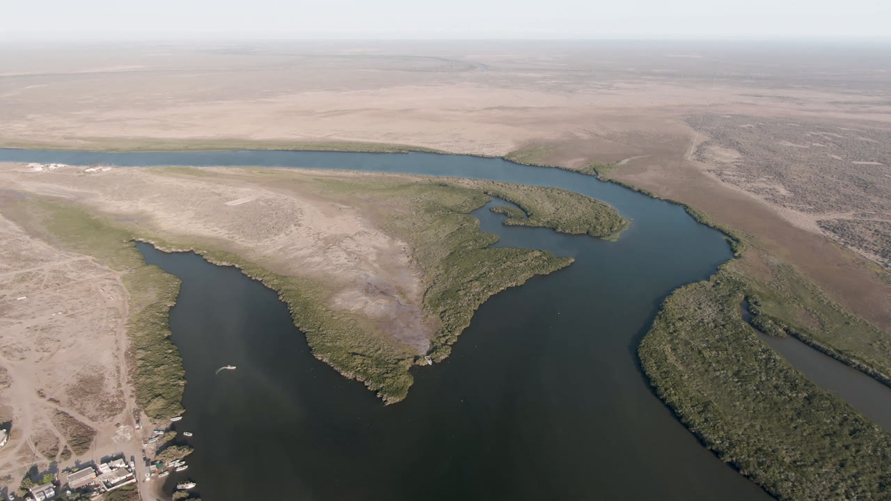 Opening aerial frame over Aldofo Lopez Mateos, Baja California Sur, with a dark winding channel between pale desert banks, scrub-covered islands, distant haze, and no visible people.