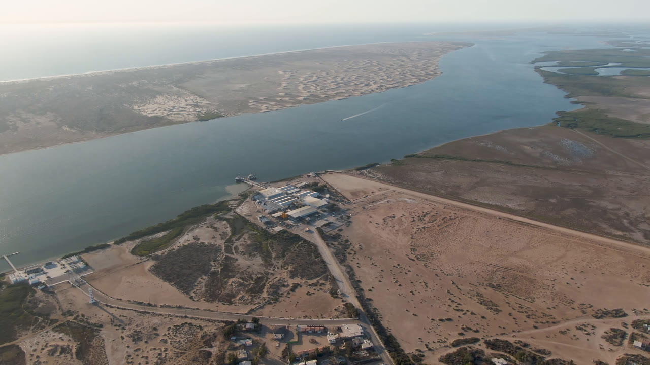 Opening aerial frame over Aldofo Lopez Mateos, Baja California Sur, showing a wide blue-green water channel, sandy foreground terrain, shoreline buildings, distant town blocks, and no visible people.