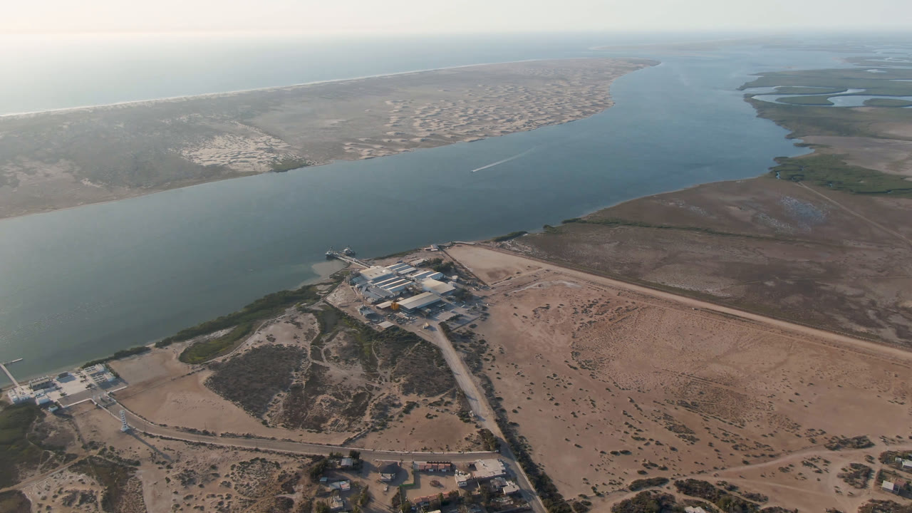 Second aerial frame over Aldofo Lopez Mateos with the waterfront complex lower in frame, the broad waterway stretching toward the horizon, distant town blocks, dry sand flats, and no visible people.