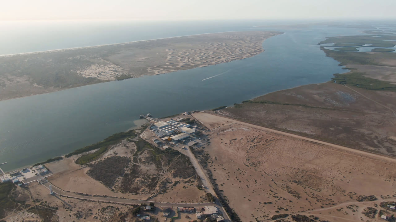 Aerial coastal landscape with water channel, sandy shoreline, buildings, and distant settlement.