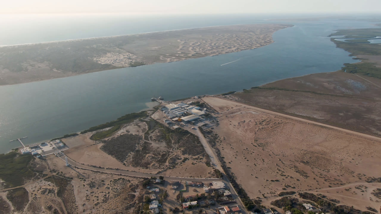 High aerial frame over Aldofo Lopez Mateos, Baja California Sur, showing blue-green water, sandy foreground, scattered buildings near a dock or bridge, and no visible people.
