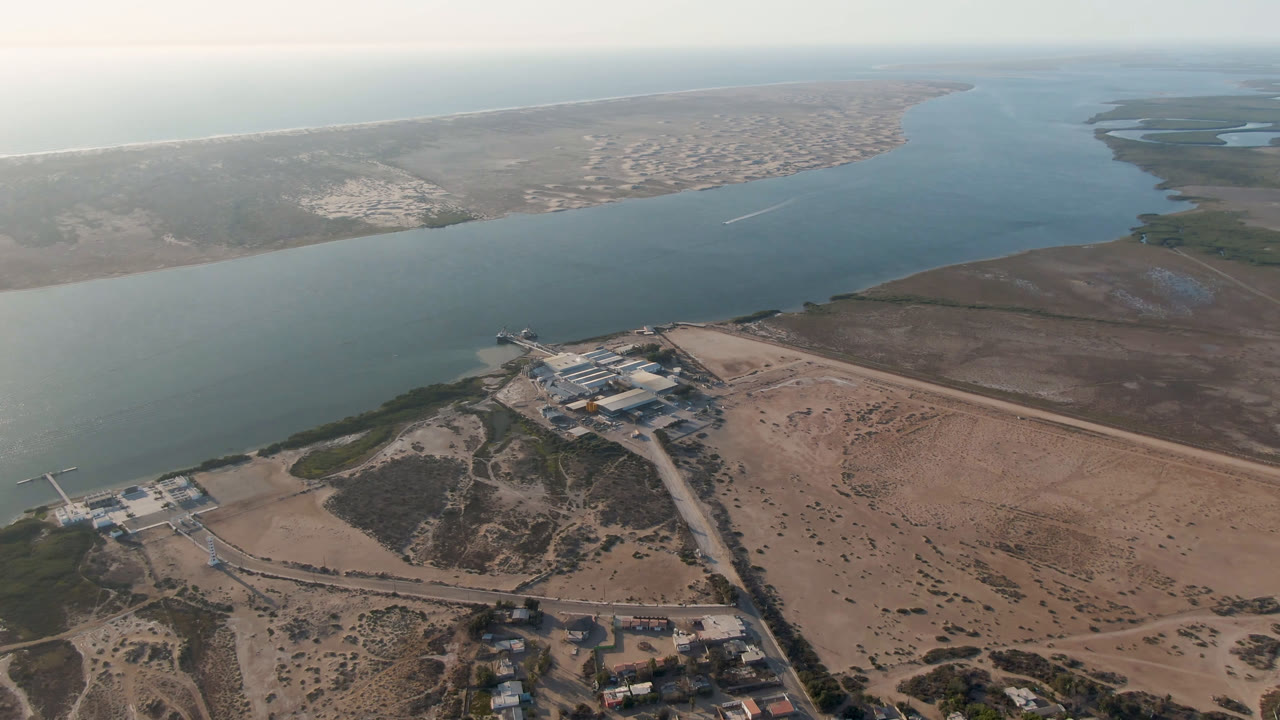 Second aerial frame in Aldofo Lopez Mateos with the water channel centered, sand and scrub in the foreground, buildings by the shore, and no visible people.