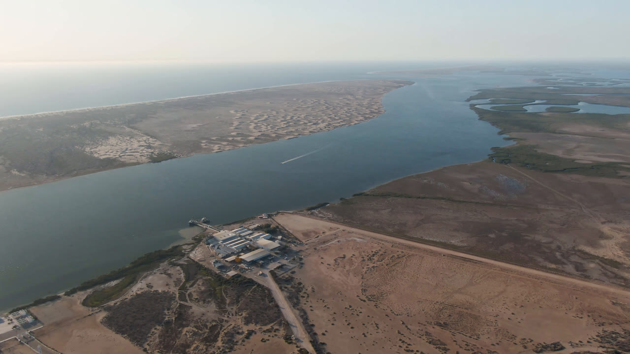 Aerial frame over Adolfo Lopez Mateos, Mexico, showing a processing plant on sandy land beside broad river water, distant coastline, and no visible people.