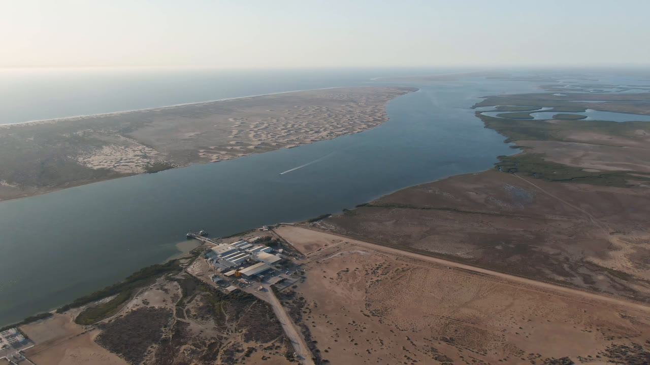Aerial view in Adolfo Lopez Mateos, Mexico, with industrial buildings near the lower frame, a wide river channel through the center, wetlands beyond, and no visible people.