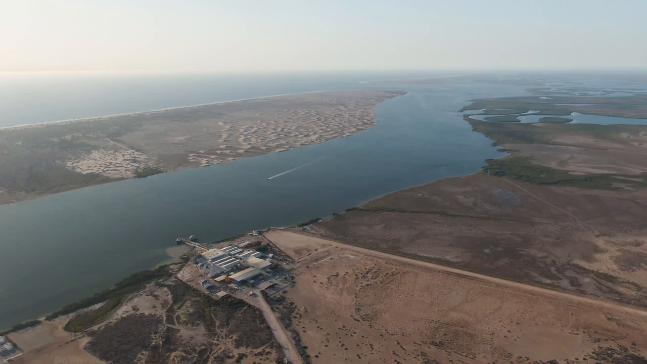 Processing facility on sandy land with river, mangroves, and ocean beyond.