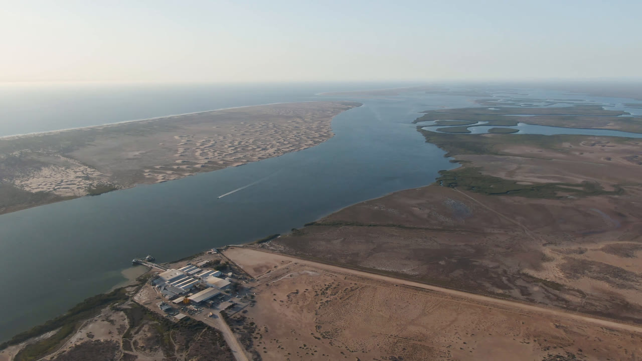 Opening aerial frame over Aldofo Lopez Mateos, Baja California Sur, showing a broad blue-green waterway between sandy shoreline and brown tidal flats, with no visible people.