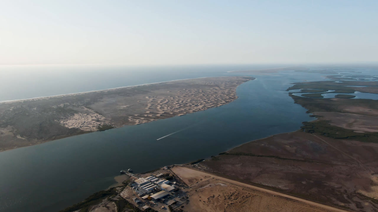 Opening aerial frame over Aldofo Lopez Mateos, Baja California Sur, with blue-green water between sandy shore and brown wetlands, hazy horizon, and no visible people.