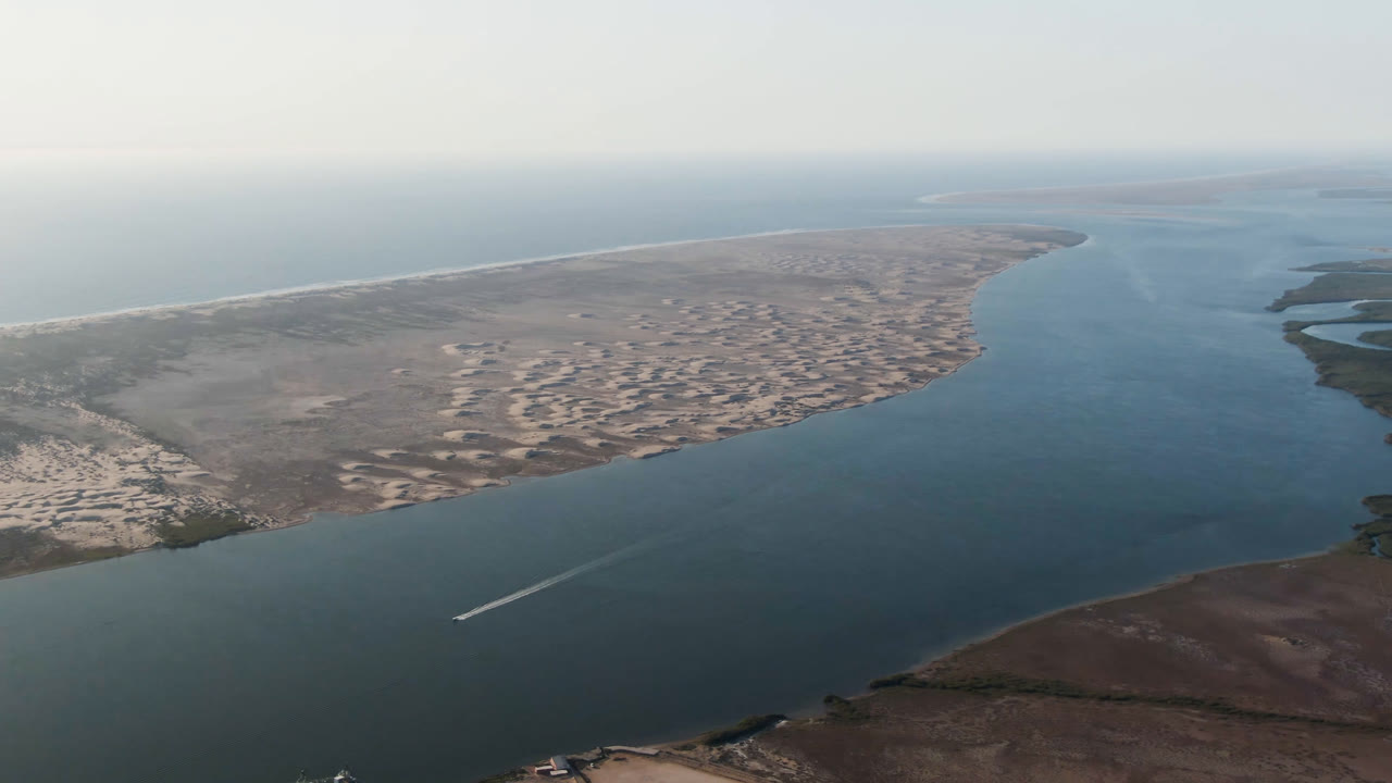 Opening aerial frame over Aldofo Lopez Mateos, Baja California Sur, with blue-green water, pale sand banks, distant ocean horizon, a small boat wake near the lower left, and no visible people.
