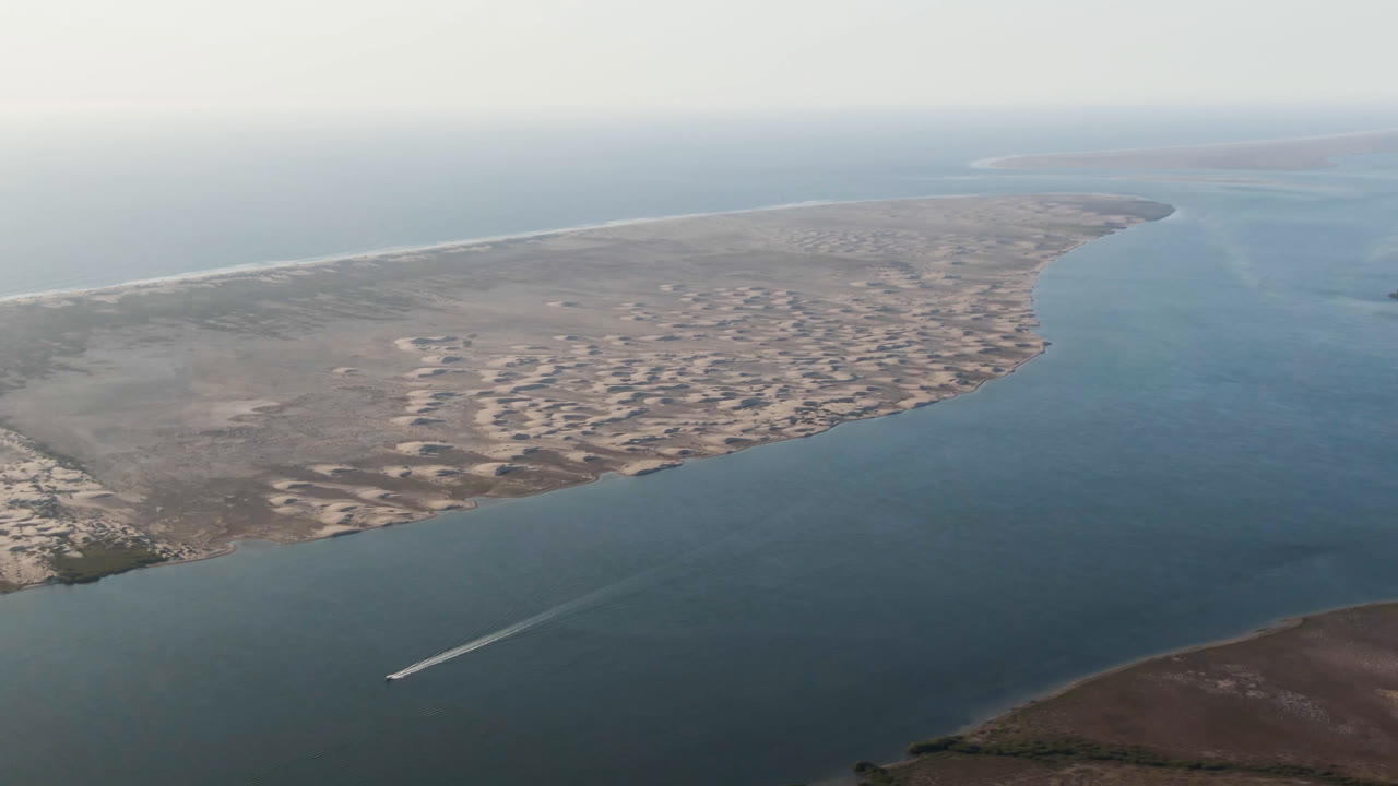 Wide drone view of coastal water, sand banks, scrubland, and a small boat wake.