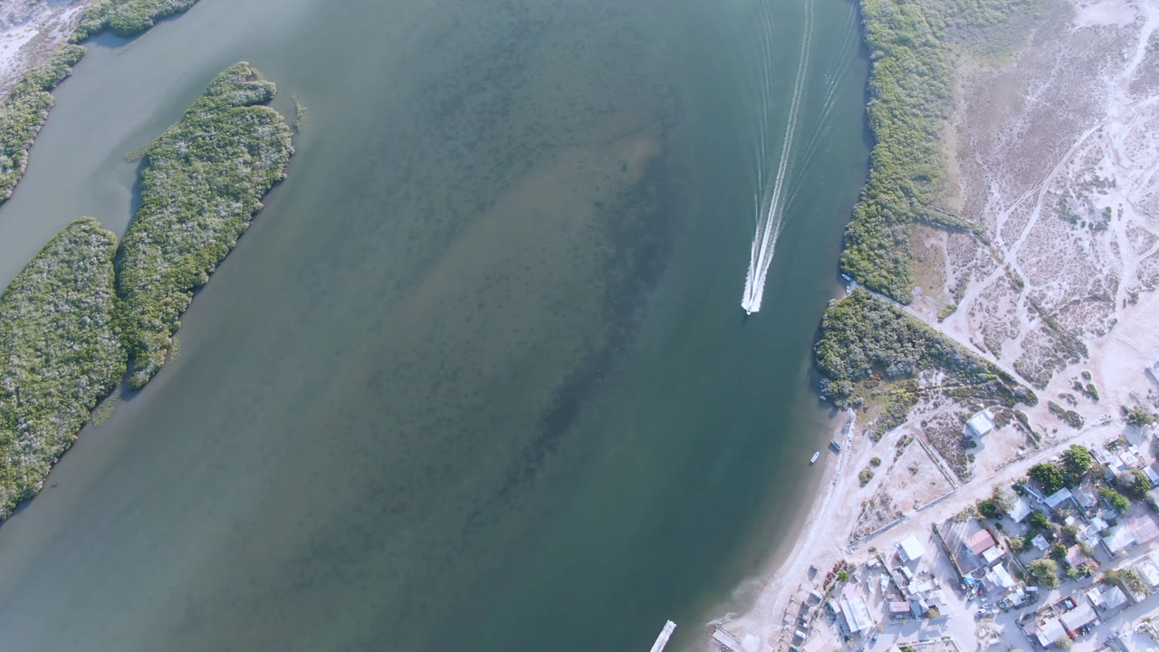 Aldofo Lopez Mateos aerial view with a small white boat farther along the channel, paired wake lines behind it, pale sand, low vegetation, dirt roads, and no visible people.