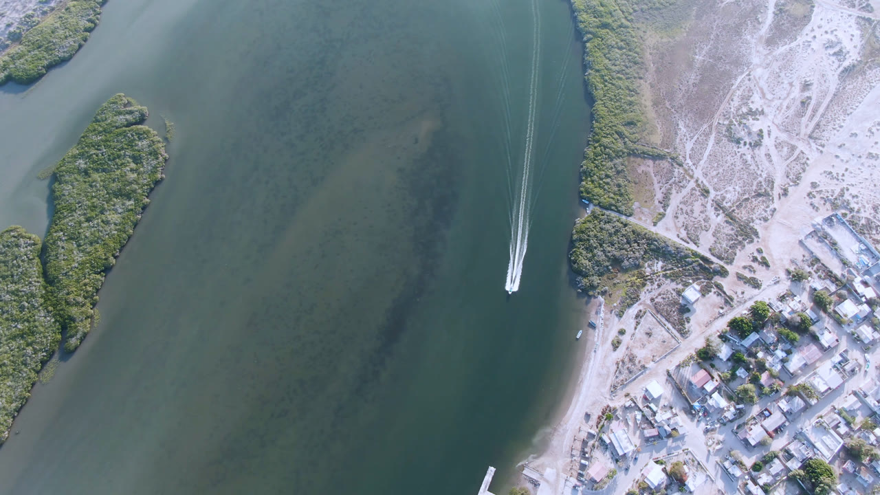 Overhead view of a boat leaving a white wake beside shoreline buildings and green water.