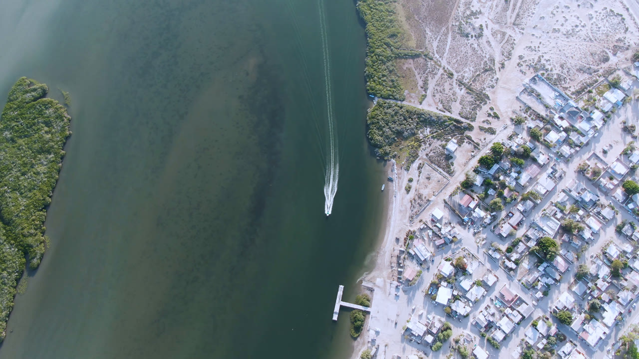 Aerial frame in Aldofo Lopez Mateos, Baja California Sur, Mexico, with the boat slightly farther along the green water, a thin wake behind it, clustered buildings to the right, and no visible people.