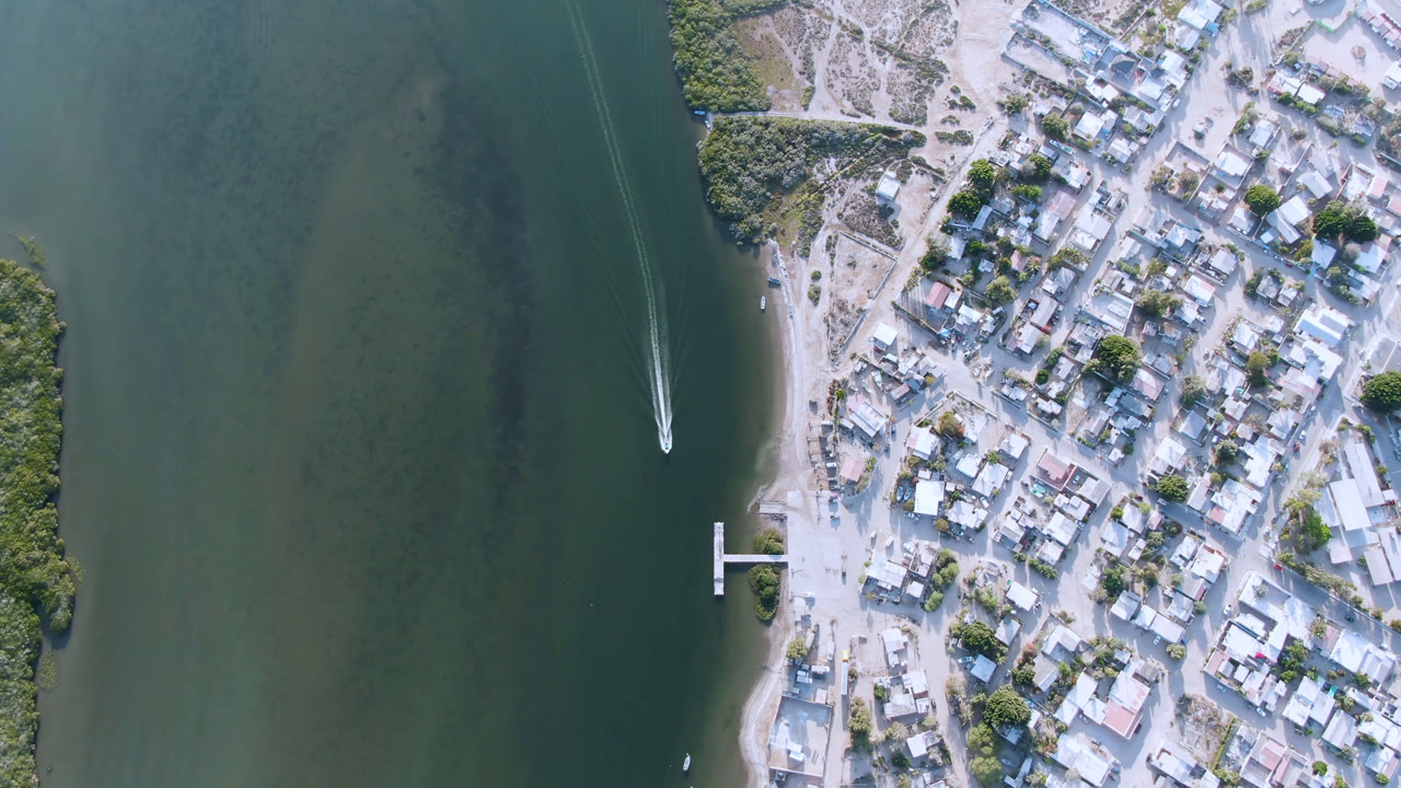 Aldofo Lopez Mateos appears from directly above with a small dock on the shoreline, compact town blocks to the right, green water to the left, and the boat wake farther along the channel, no visible people.