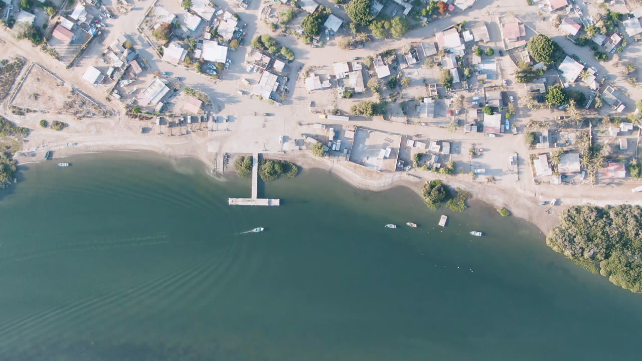 Aerial view of Aldofo Lopez Mateos showing the pier near center, a small boat leaving a faint wake, several other boats near shore, pale roads, low buildings, and no visible people.