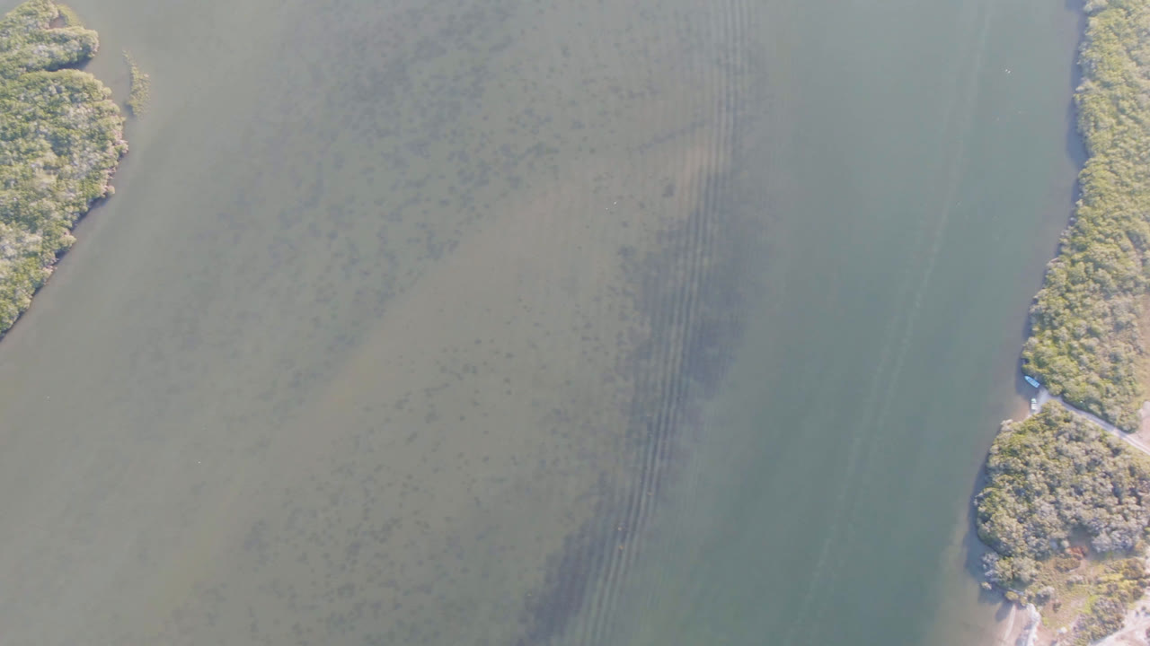 A narrow vegetated island sits between pale water channels in Aldofo Lopez Mateos, with faint underwater bands and no visible people.