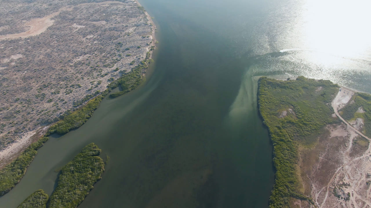 High aerial frame over Aldofo Lopez Mateos, Baja California Sur, showing dark river channels, dry scrub islands, green shoreline vegetation, bright water glare, and no visible people.