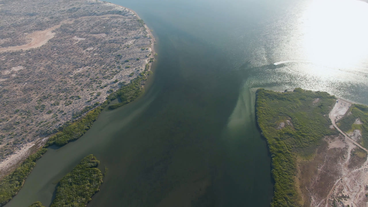 Overhead view in Aldofo Lopez Mateos, Baja California Sur, with sun glare spreading across the river, scrubland on both sides, a small white boat wake on the water, and no visible people.
