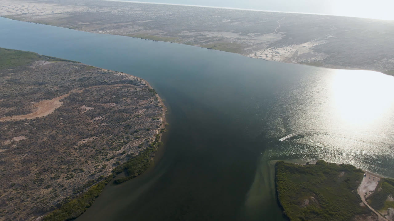 High aerial view over Aldofo Lopez Mateos, Baja California Sur, with a dark water channel curving around scrub and sand, bright sun reflection on the surface, a small boat wake near the lower center, and no visible people.