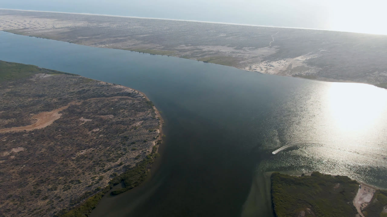 Aerial frame from Aldofo Lopez Mateos, Baja California Sur, showing the river delta from above with scrubland in the foreground, bright white glare on the water, pale shoreline in the distance, and no visible people.