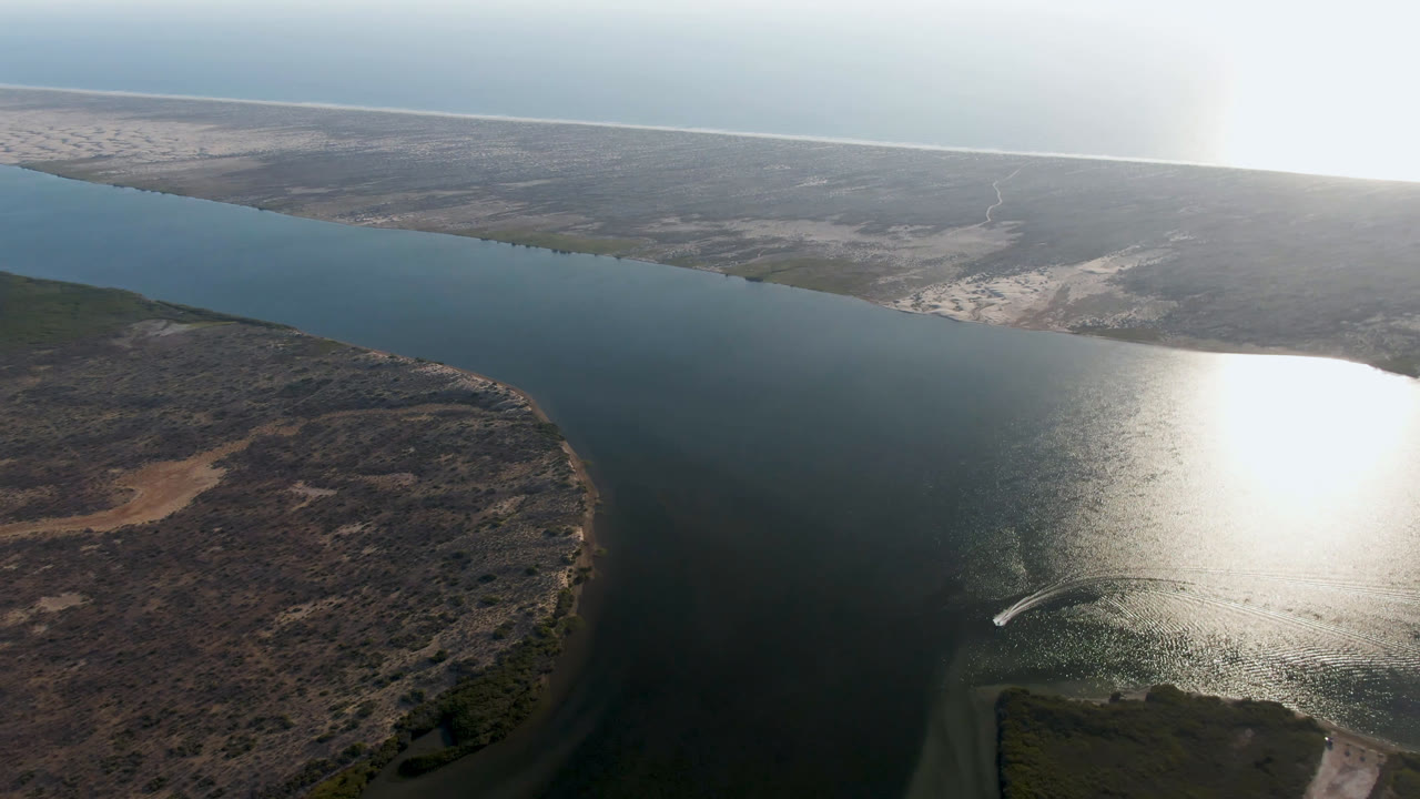Oblique drone view of water, low scrubland, sand, horizon, and a curved wake near the glare.
