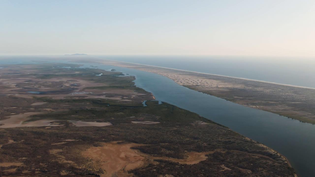 Wide aerial frame over Aldofo Lopez Mateos, Baja California Sur, with a broad water channel, dry scrubland, pale sandbar, distant ocean horizon, and no visible people.