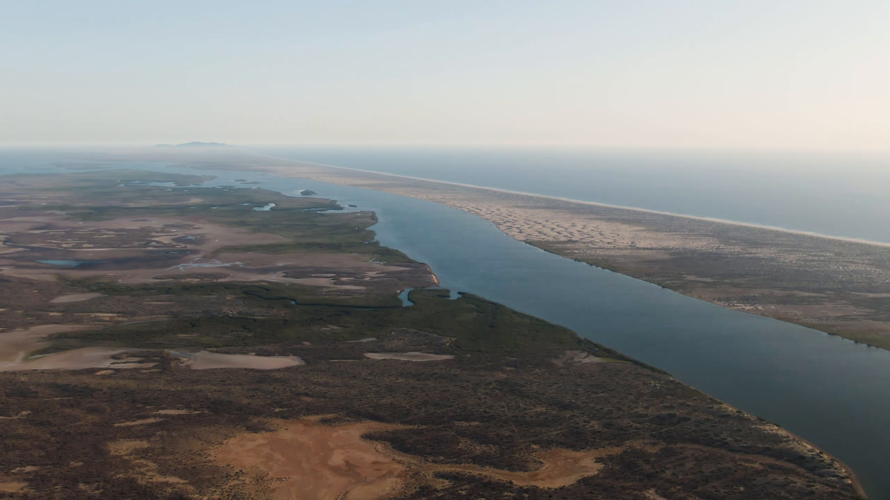 A high aerial view in Aldofo Lopez Mateos, Baja California Sur, shows blue water widening through dry brown terrain toward a long pale beach and ocean, with no visible people.