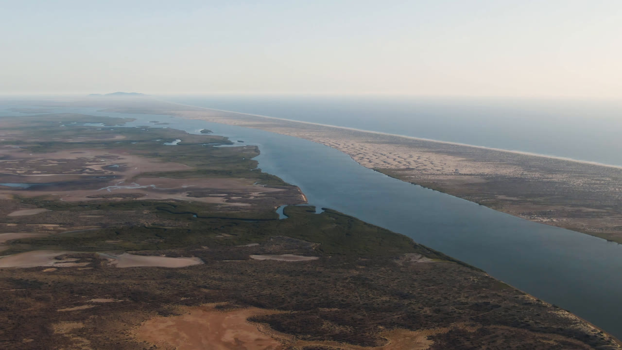 Aerial frame over Aldofo Lopez Mateos, Baja California Sur, with the channel angled through dark scrub and tan flats, a pale beach strip on the right, ocean beyond, and no visible people.