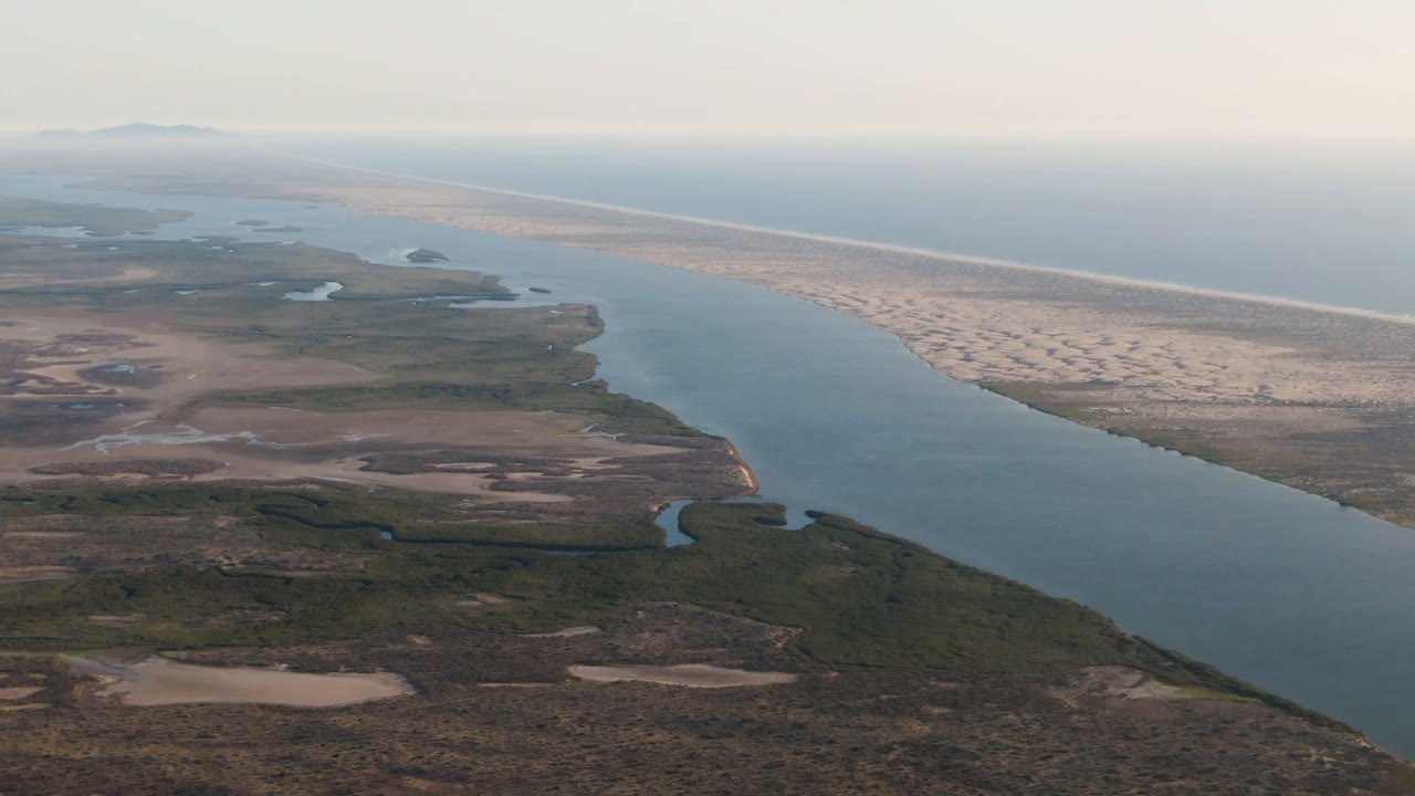 Opening aerial frame over Aldofo Lopez Mateos, Baja California Sur, showing a broad water channel, desert scrub, pale sandbar, ocean horizon, and no visible people.