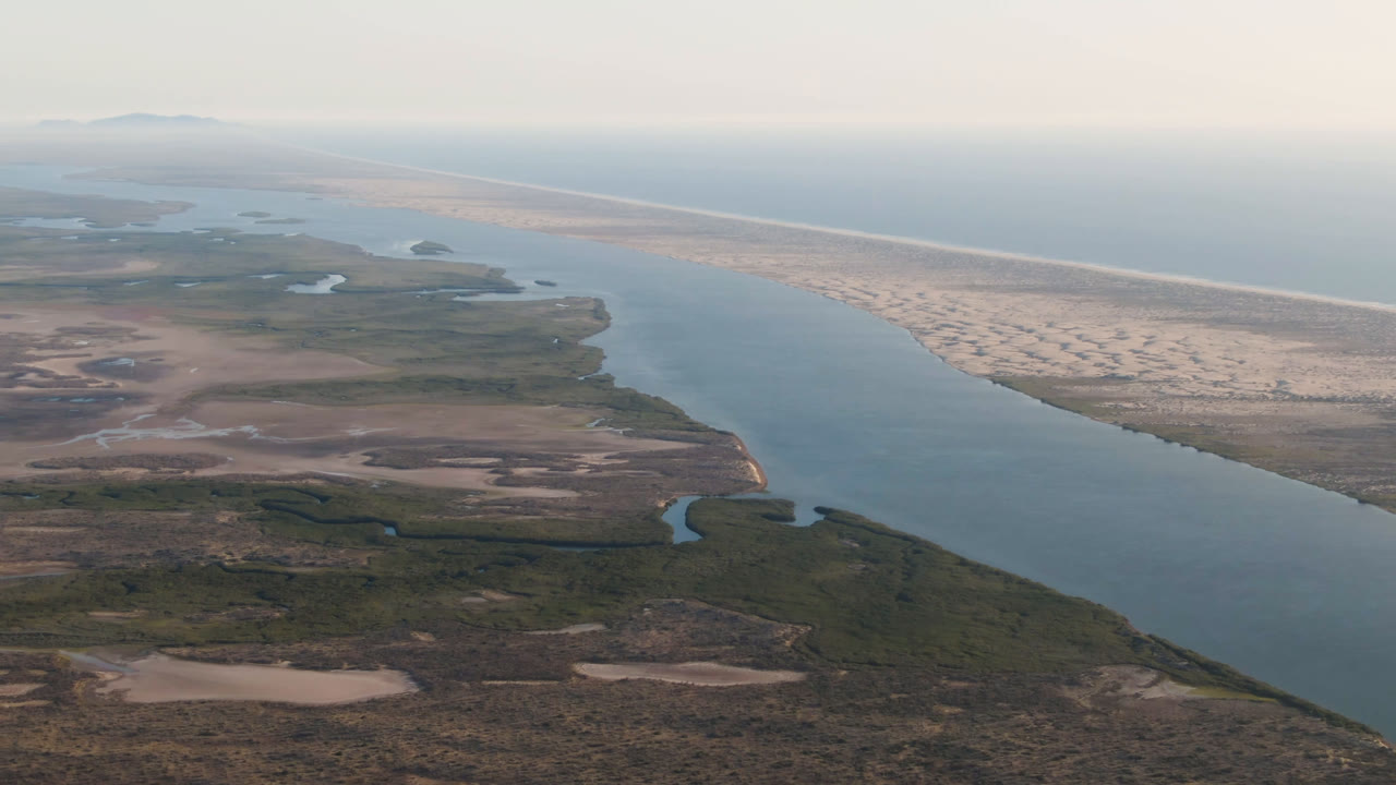 Second aerial frame in Aldofo Lopez Mateos, Baja California Sur, with the main blue channel centered, scrubland in the foreground, a pale sand strip beyond it, and no visible people.