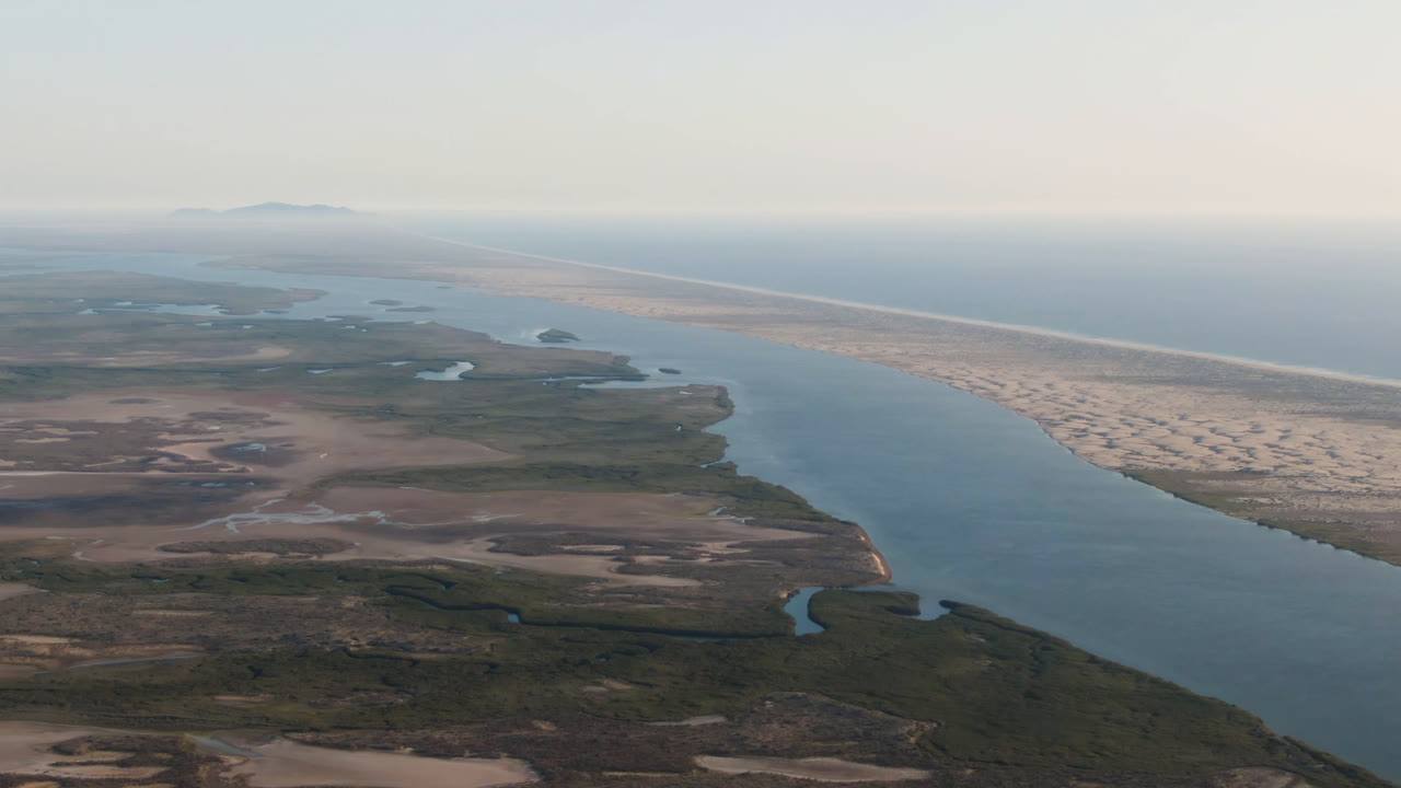 Wide aerial frame over Aldofo Lopez Mateos, Baja California Sur, showing a broad river channel, sandy coastal strip, ocean horizon, scrubland foreground, and no visible people.