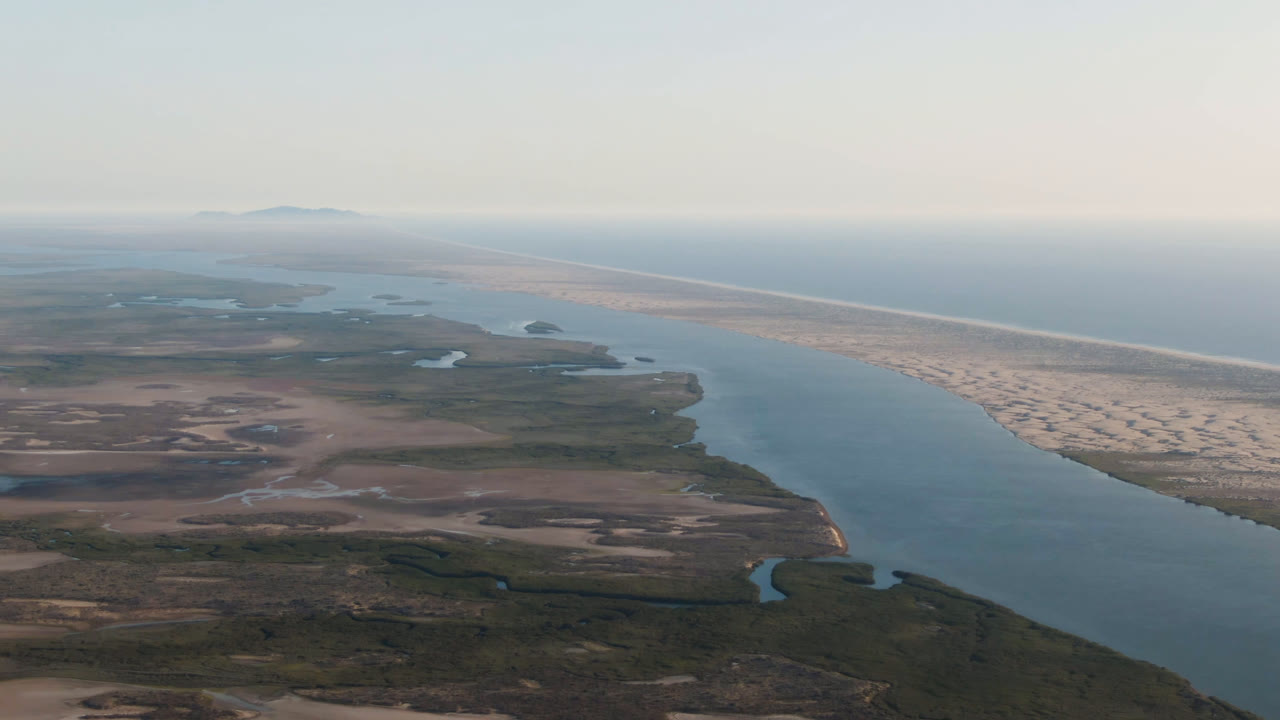 Aerial frame in Aldofo Lopez Mateos with the river channel centered, arid flats and winding inlets below, a pale beach strip beyond, and no visible people.