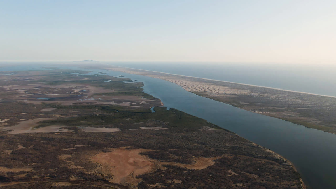 Wide aerial frame over Aldofo Lopez Mateos, Baja California Sur, with a blue river cutting past dark desert vegetation toward a pale beach and ocean horizon, no visible people.