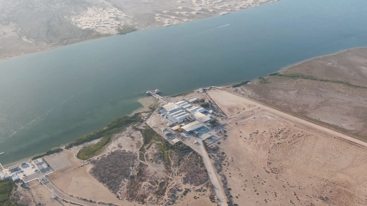 Opening aerial frame over Aldofo Lopez Mateos, Baja California Sur, with blue-green water, pale shore, roads, low buildings, distant settlement, and no visible people.
