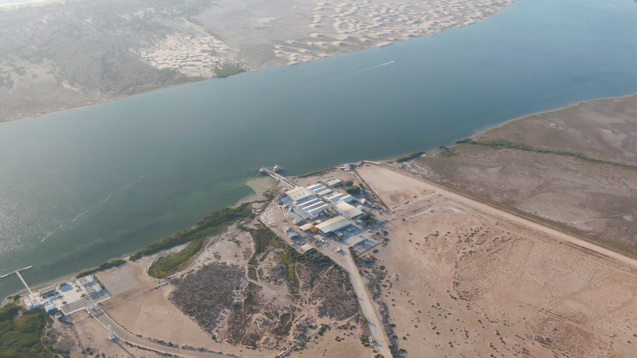 Aerial frame in Aldofo Lopez Mateos showing the waterway across the center, waterfront structures lower in frame, dry coastal terrain, distant houses, and no visible people.