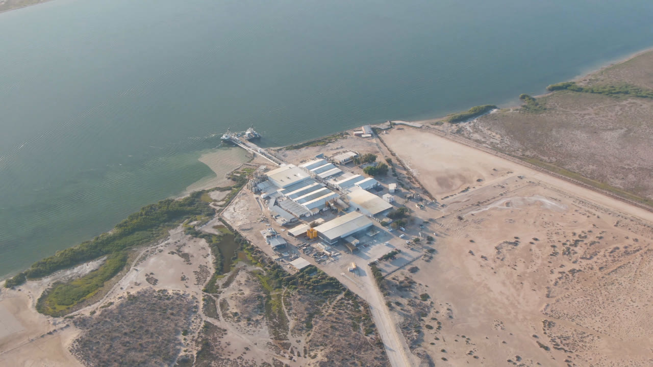 Aerial frame over Aldofo Lopez Mateos, Baja California Sur, showing a pale-roofed building complex near the shoreline, sand tracks, scrub, and green-blue water with no visible people.