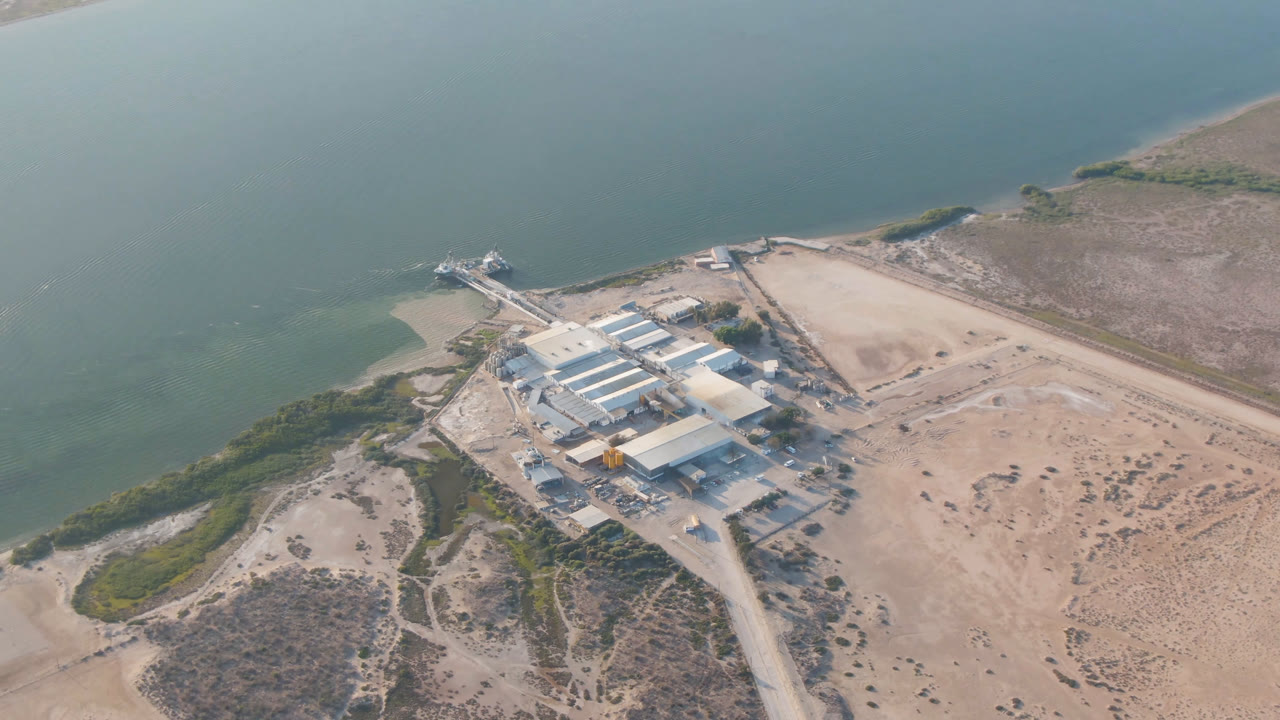 Aerial view in Aldofo Lopez Mateos, Baja California Sur, with the building complex centered below the water, a road entering from the lower frame, shoreline scrub, and no visible people.
