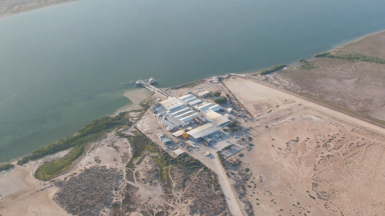 Drone view of a coastal building complex with pale roofs, sand flats, scrub, and water behind it.