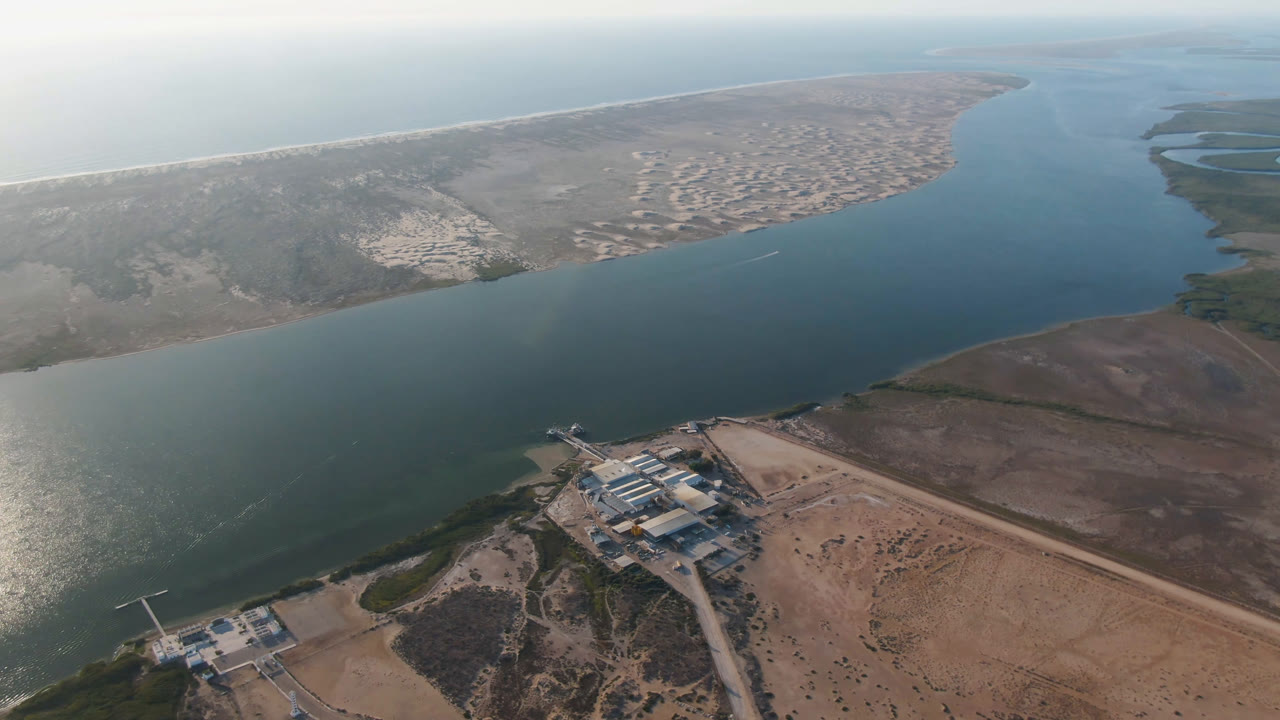 Aerial frame over Aldofo Lopez Mateos, Baja California Sur, showing a wide blue-green channel, sandy barrier land, ocean surf beyond, a small shoreline compound below, and no visible people.