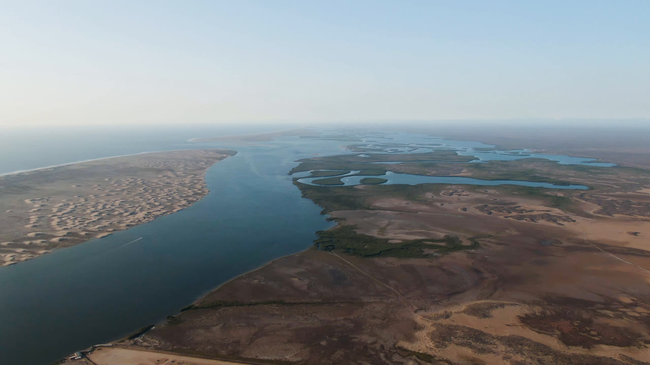 Opening aerial frame over Aldofo Lopez Mateos, Baja California Sur, with a wide dark river between sandy ocean shoreline and dry desert flats, no visible people.