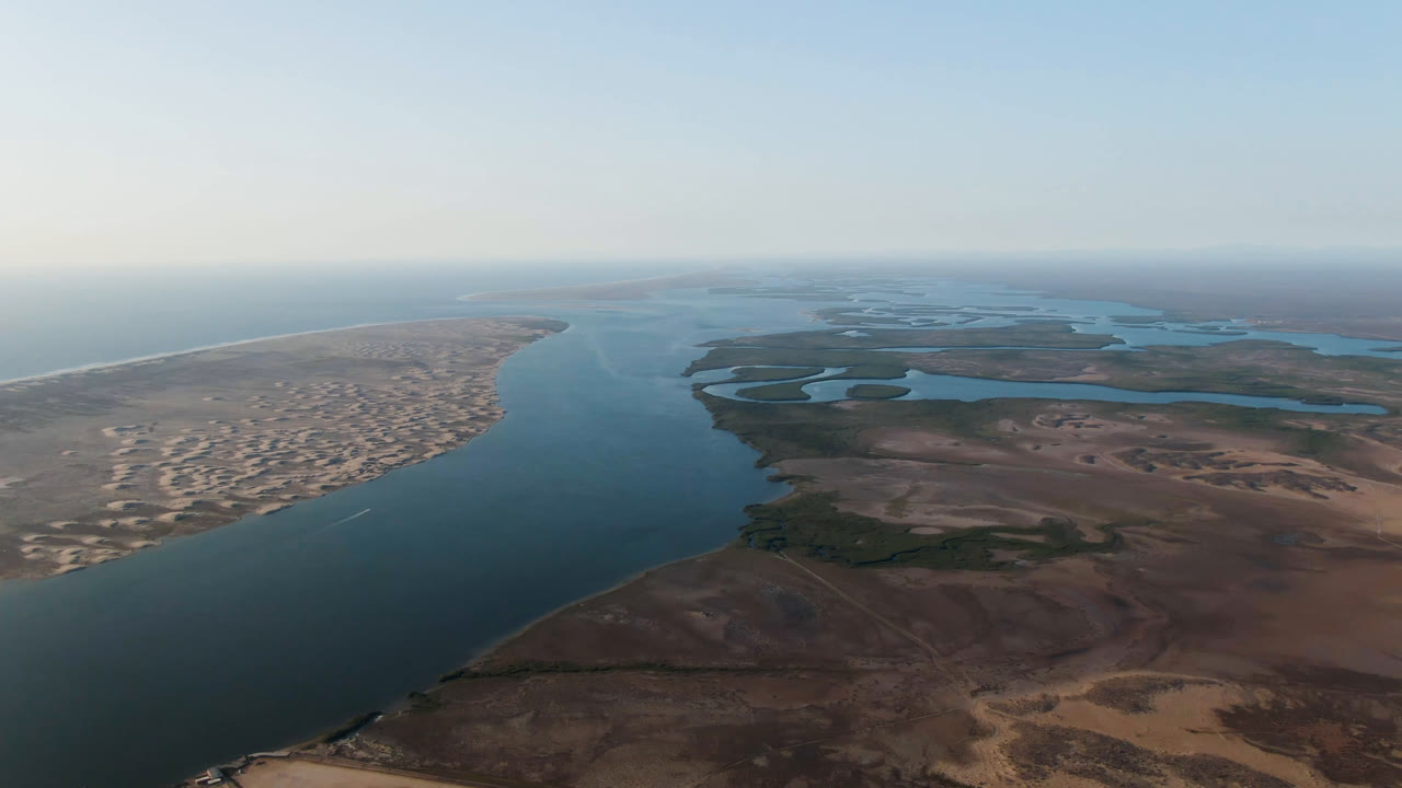 Wide aerial view of a coastal waterway, sandy shore, brown flats, and pale horizon.