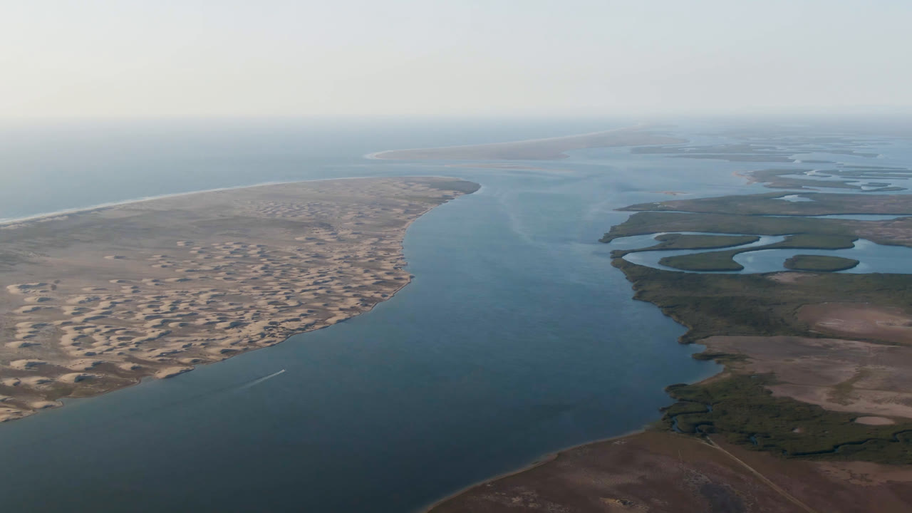 High aerial frame over Aldofo Lopez Mateos, Baja California Sur, showing a wide blue channel, pale sand dunes to the left, dark marshland to the right, hazy horizon, and no visible people.