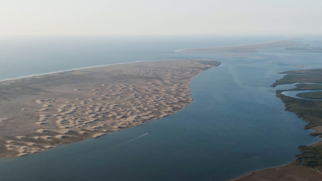 Wide aerial frame over Aldofo Lopez Mateos, Baja California Sur, with a blue water channel, pale sand dunes on the left, low shoreline on the right, hazy sky, and no visible people.