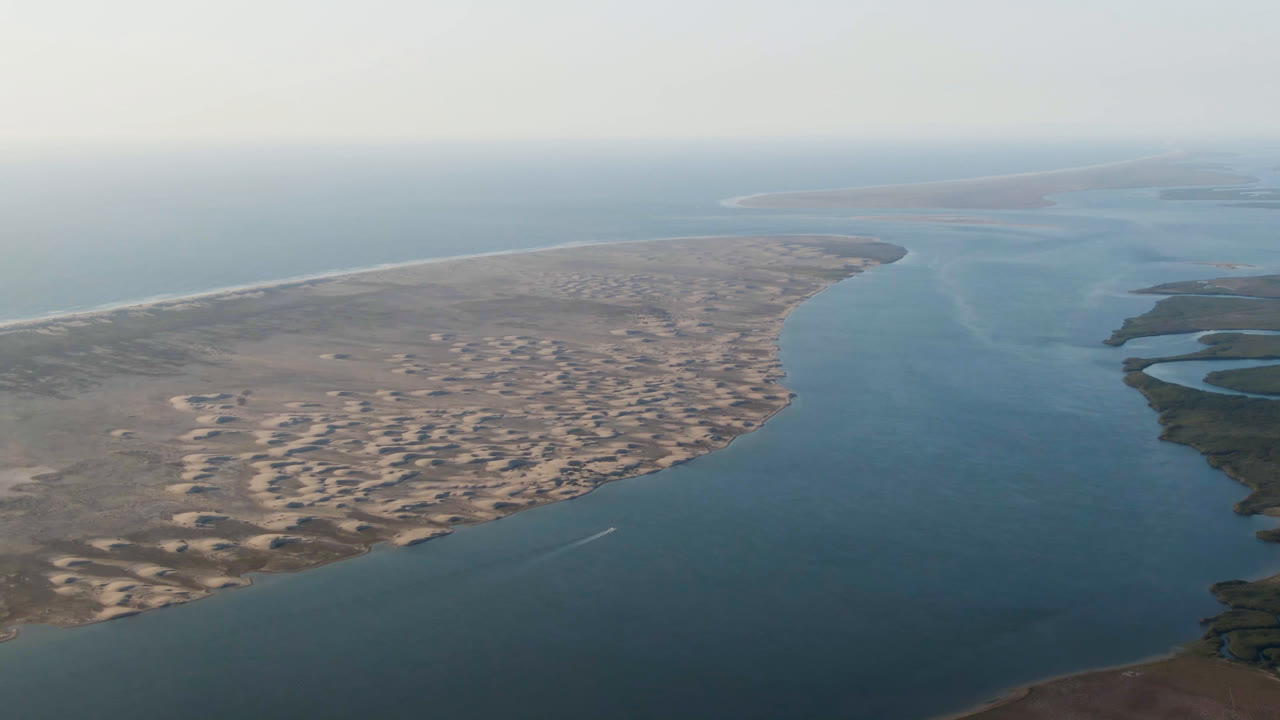 Aerial view at Aldofo Lopez Mateos showing the waterway curving past a broad dune field, with darker wetland edges to the right, distant ocean haze, and no visible people.