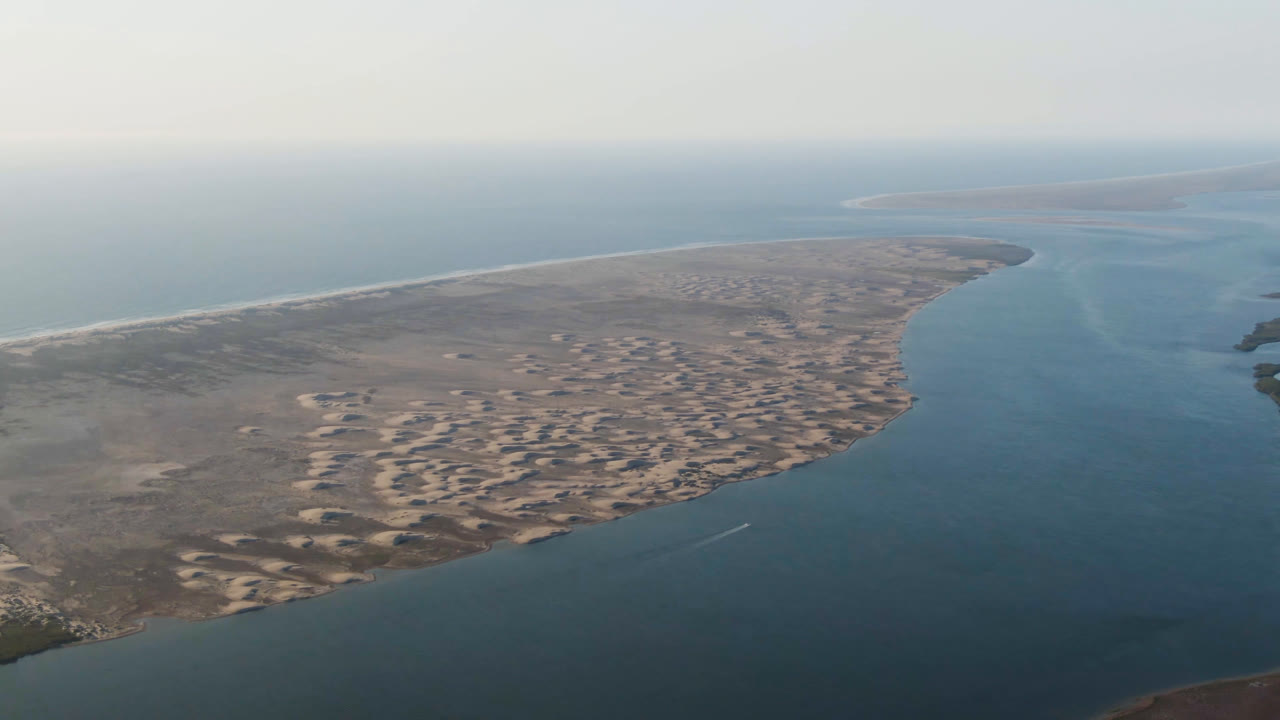 Opening aerial frame over Aldofo Lopez Mateos, Baja California Sur, showing pale sandy terrain, a broad blue water channel, distant ocean, and no visible people.
