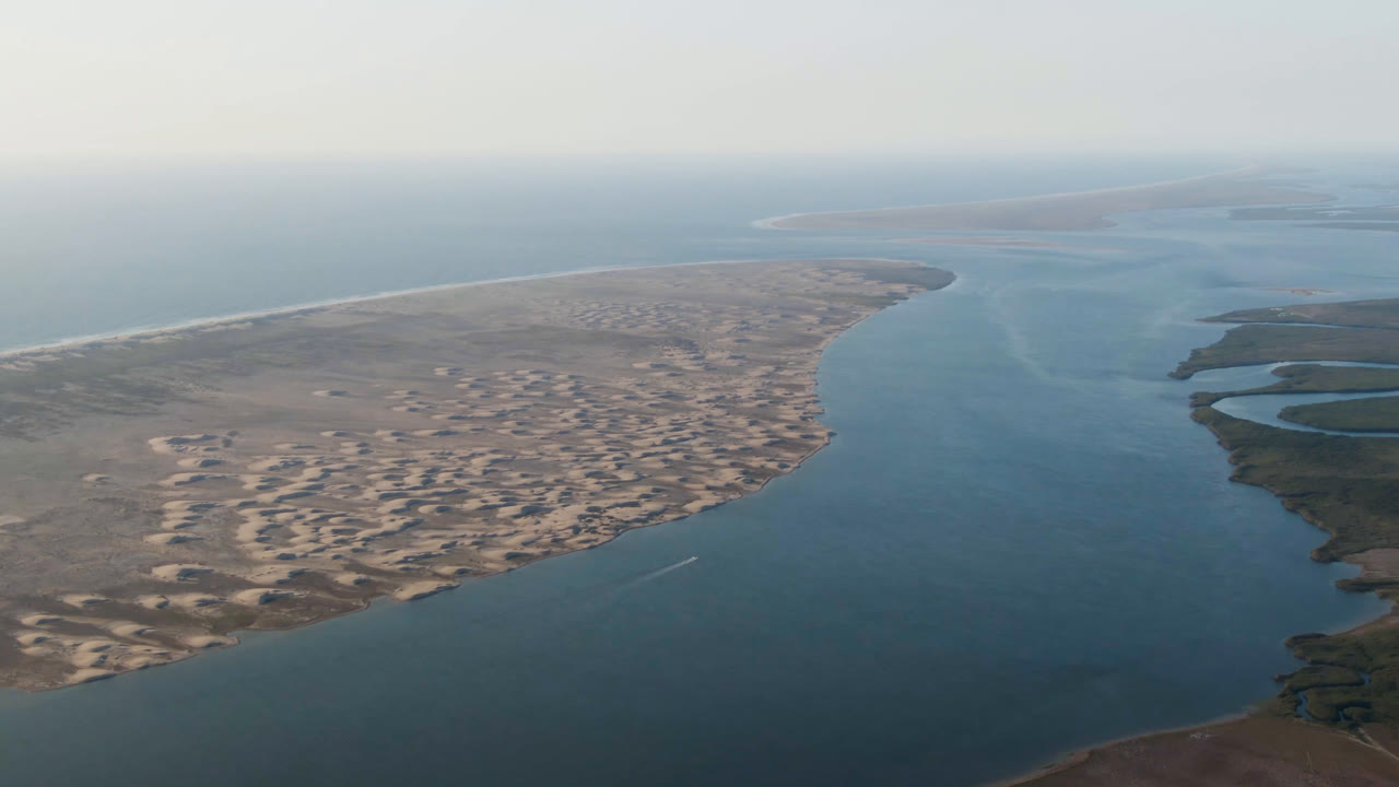 High aerial view over Aldofo Lopez Mateos, Baja California Sur, with blue water in the foreground, pale dunes along the shore, a hazy horizon, and no visible people.