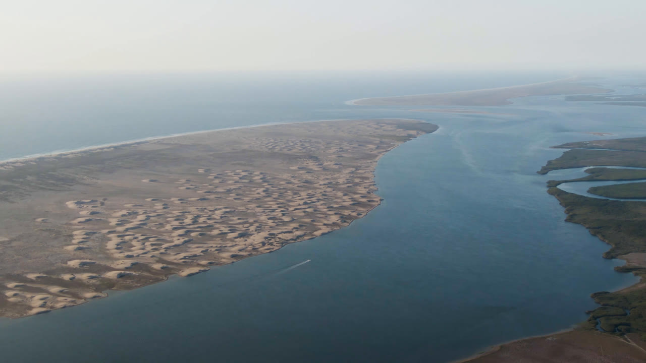 Opening aerial frame over Aldofo Lopez Mateos, Baja California Sur, Mexico, with broad blue water around pale sand banks, low dunes, a hazy horizon, and no visible people.