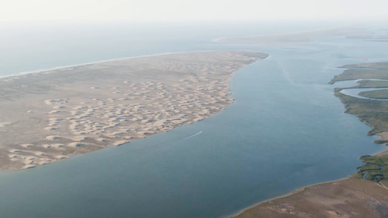 High aerial frame from Aldofo Lopez Mateos, Baja California Sur, showing pale sand and low scrub beside broad calm water, with the horizon softened by haze and no visible people.