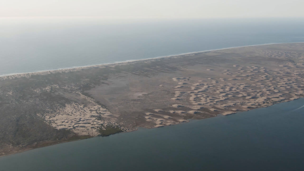 Aerial frame from Aldofo Lopez Mateos, Baja California Sur, looking across dark water to a sandy shoreline, scrubby sand banks, distant surf, and no visible people.